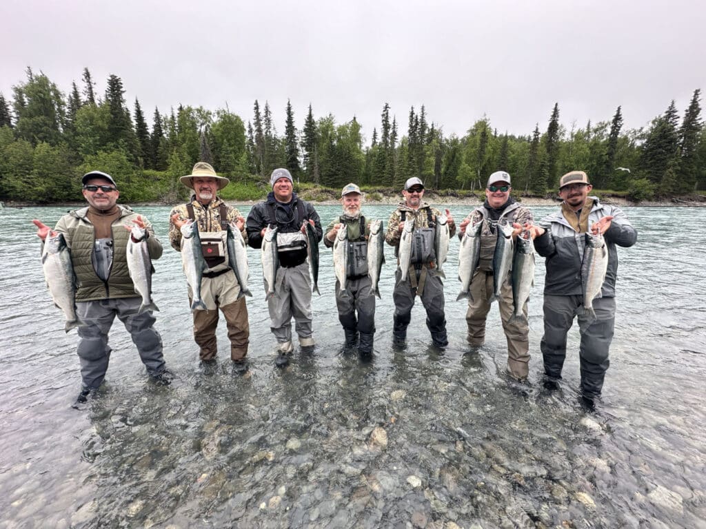 A group of seven anglers standing waist‑deep in the clear, gravel‑bottomed waters of the Kasilof River, each holding two large silver salmon. They’re dressed in waders and waterproof gear, with spruce and birch trees lining the opposite riverbank under an overcast sky.